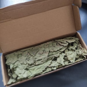 Peut inclure: Une boîte en carton avec le couvercle ouvert, révélant un paquet de feuilles vertes séchées.