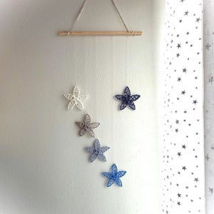 Puede incluir: Un móvil con cinco estrellas de color blanco, gris y azul en forma de estrella que cuelgan de una varilla de madera. El móvil está suspendido del techo.