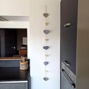 Pode incluir: Uma decoração de parede vertical composta por nove ornamentos em forma de coração, alternando entre cinza e creme, enfiados num fio fino. Os corações são texturizados e dispostos numa única coluna, suspensos por um pequeno laço no topo.