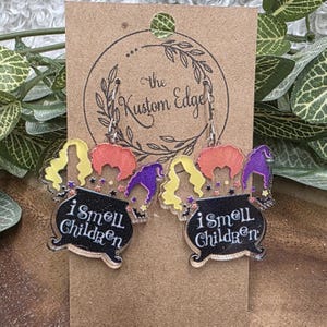 May include: A pair of dangle earrings featuring a black cauldron with the text "i smell children". Above the cauldron are colorful cartoon wigs in yellow, orange, and purple. The earrings are on a brown card with the text "The Kustom Edge".