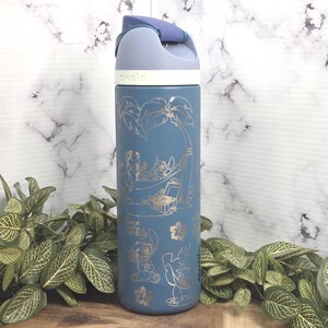May include: A blue insulated water bottle with a flip-top lid. The bottle features gold-colored illustrations of characters and tropical elements. The word "awala" is printed on the lid. The bottle is surrounded by green plants.