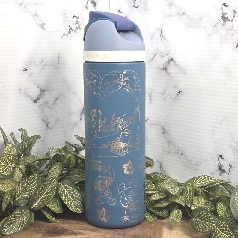 May include: A blue insulated water bottle with a flip-top lid. The bottle features gold-colored illustrations of characters and tropical elements. The word "awala" is printed on the lid. The bottle is surrounded by green plants.