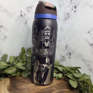 Puede incluir: Una botella de agua aislada azul oscuro con un dise&ntilde;o plateado grabado con villanos de Disney. La botella tiene una tapa negra con una banda azul y un asa marr&oacute;n. El dise&ntilde;o incluye im&aacute;genes de la Reina Malvada y otros personajes.