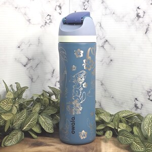 May include: A blue insulated water bottle with a flip-top lid. The bottle features a silver floral and character design. The word "owala." is printed on the side. The lid is light blue and purple.