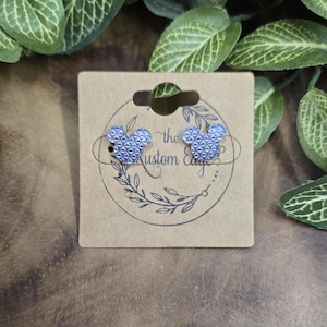 May include: A pair of blue Mickey Mouse-shaped stud earrings with a geometric pattern, displayed on a brown card. The card has the text "the Kustom Edge" and a floral design. The background includes green leaves.