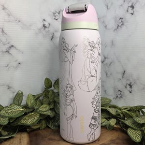 Pode incluir: Uma garrafa de &aacute;gua Owala rosa claro com desenhos de linha de princesas da Disney. A garrafa tem uma al&ccedil;a cinza e uma tampa rosa e verde claro. O logotipo da Owala &eacute; impresso em dourado na lateral da garrafa.