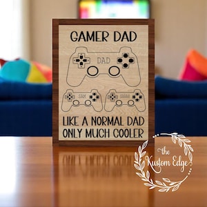 Puede incluir: Cartel enmarcado de madera con el texto "GAMER DAD" y "LIKE A NORMAL DAD ONLY MUCH COOLER". El cartel presenta gráficos de mando y los nombres "SAM" y "SARAH". El cartel está sobre una mesa de madera.