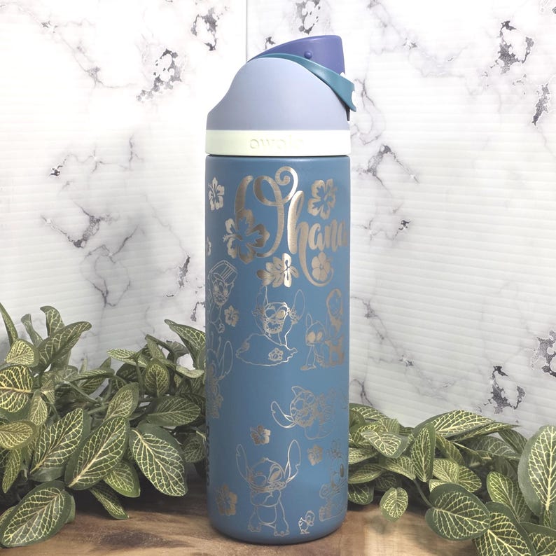 May include: A blue water bottle with a flip-top lid. The bottle features a silver design with the word "Ohana" and character illustrations. The lid is light blue and purple. The bottle is surrounded by green plants.