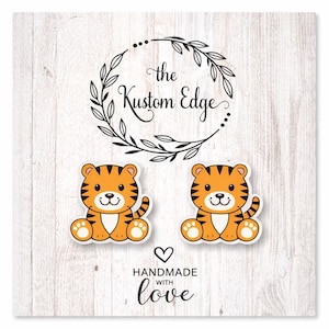 May include: Two orange and black tiger-shaped earrings with white accents. The earrings are on a white background with the text "the Kustom Edge" and "HANDMADE with love". The earrings have a cute cartoon design.