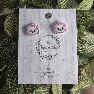 May include: A pair of white and pink cat-shaped stud earrings. The earrings are displayed on a white card with the text "the Kustom Edge" and "HANDMADE with love". The earrings have pink bows and are set against a backdrop of green leaves.