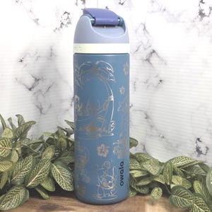 May include: Blue insulated water bottle with a flip-top lid. The bottle features a silver design of a cartoon character and tropical elements. The brand name "owala" is printed on the side. The bottle is surrounded by green plants.