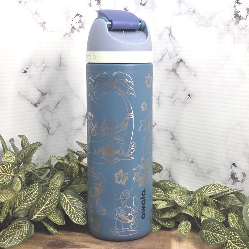May include: Blue insulated water bottle with a flip-top lid. The bottle features a silver design of a cartoon character and tropical elements. The brand name "owala" is printed on the side. The bottle is surrounded by green plants.