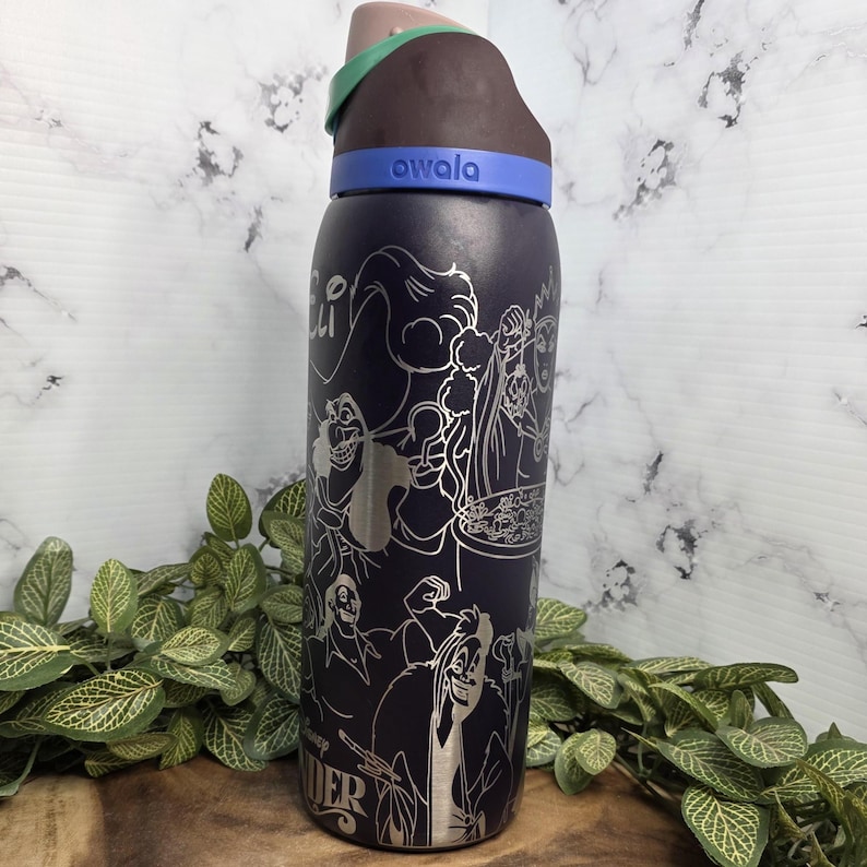 Puede incluir: Una botella de agua Owala azul oscuro con un dise&ntilde;o plateado grabado de villanos de Disney. La botella tiene una tapa marr&oacute;n, verde y azul. La palabra "owala" est&aacute; impresa en la banda azul.