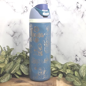 May include: A blue water bottle with a flip-top lid. The bottle is decorated with gold-colored graphics of characters and the word "Ohana". The lid is light blue and has a push-button release. The bottle is surrounded by green plants.