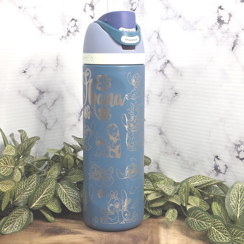 May include: A blue water bottle with a flip-top lid. The bottle is decorated with gold-colored graphics of characters and the word "Ohana". The lid is light blue and has a push-button release. The bottle is surrounded by green plants.
