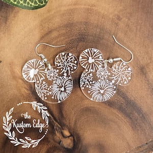 Puede incluir: Un par de pendientes de acrílico transparente con forma de orejas de Mickey Mouse con un diseño de fuegos artificiales blancos. Los pendientes están sobre una superficie de madera.