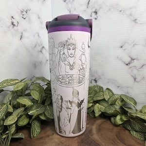 Puede incluir: Un termo de viaje rosa claro con tapa negra y morada. El termo presenta un dibujo lineal plateado de villanos de Disney. El termo est&aacute; rodeado de follaje verde.