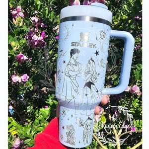 May include: A light blue Stanley tumbler with a handle, featuring a design of a prince and princess, stars, and other illustrations. The tumbler has a white lid and a light blue handle. The brand name "Stanley" is visible on the front.