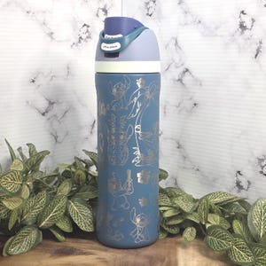 May include: A blue water bottle with a light blue and purple lid. The bottle features gold-colored illustrations of a cartoon character and tropical elements. The lid has a push button and the text "Lift to Unlock".