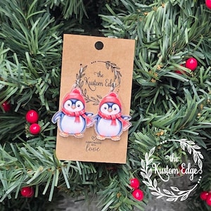 May include: A pair of penguin earrings with red hats and scarves, displayed on a brown tag with the text "the Kustom Edge" and "handmade with love". The earrings are set against a backdrop of green pine branches and red berries.
