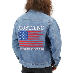 Op de afbeelding: Een blauwe denimjas met de tekst "MUSTANG" en "AMERICA'S PONY CAR" in wit op de rug. Een rood, wit en blauwe Amerikaanse vlag grafiek is gedrukt op de rug van de jas.