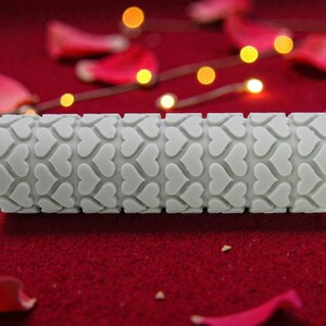 May include: A gray rolling pin with a heart pattern. The roller is cylindrical and features a repeating design of hearts. The background is a deep red surface with scattered rose petals and small lights.