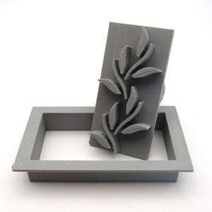 May include: A gray plastic stamp with a design of stylized leaves. The stamp is sitting in a gray plastic frame.