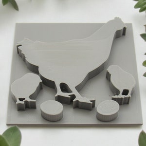 May include: A gray 3D printed design of a hen and two chicks with two eggs. The hen is standing with its wings spread out. The chicks are standing next to the hen. The eggs are round and smooth.
