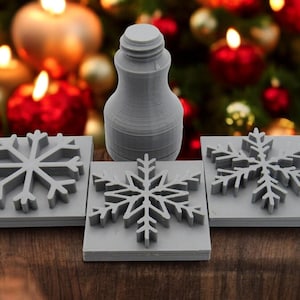 May include: Three gray snowflake-shaped rubber stamps and a gray bottle stopper. The stamps are square and have a raised snowflake design. The bottle stopper is cylindrical and has a textured surface.