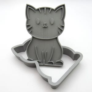 Pode incluir: Cortador de biscoitos de plástico cinza em forma de gato. O cortador tem um design detalhado com um padrão listrado e um rosto bonito.