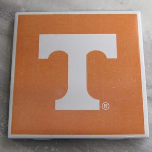 Puede incluir: Posavasos de cerámica naranja con una "T" blanca en el centro. La "T" tiene un símbolo de marca registrada a la derecha.