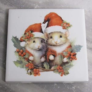 Può includere: Due topi bianchi che indossano cappelli rossi di Babbo Natale e tengono rami di agrifoglio con bacche rosse. I topi sono su uno sfondo bianco.