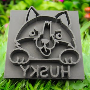 Könnte beinhalten: Ein grauer Gummistempel mit einem Cartoon-Husky-Hundedesign. Der Hund lächelt und hat die Zunge herausgestreckt. Das Wort "HUSKY" ist unter dem Hund gedruckt.