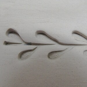 May include: A close-up of a white ceramic surface with a decorative design of a stylized branch with leaves. The design is embossed into the surface, creating a raised effect.