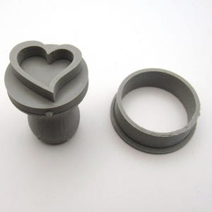 May include: A gray heart-shaped cookie stamp and a matching round cutter. The stamp features a raised heart design. The items are made of plastic and are designed for baking. The tools are on a white background.