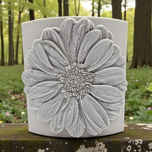 May include: A light gray cylindrical vessel featuring a large, three-dimensional sculpted flower on its front surface. The flower has numerous textured petals radiating from a detailed central disk, creating a prominent relief design on the smooth container.