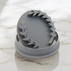 May include: A gray plastic cookie cutter in the shape of a wreath with leaves. The cutter is open and the top part is sitting on top of the bottom part.