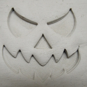 May include: A white surface with a carved jack-o'-lantern face. The face has two crescent-shaped eyes, a triangular nose, and a jagged mouth.