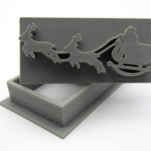 Puede incluir: Una caja de plástico gris con una tapa con bisagras. La tapa presenta un diseño en relieve de Papá Noel en un trineo tirado por renos.