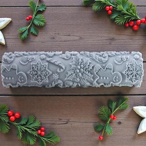 May include: A gray, textured rolling pin with embossed Christmas-themed designs, including snowflakes, gingerbread men, and holly. The image is decorated with star-shaped cookies and sprigs of greenery with red berries.