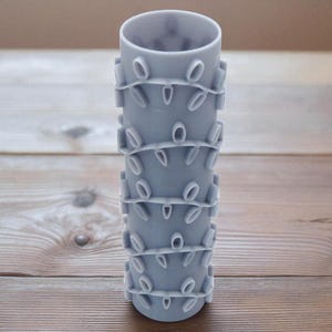 May include: A light grey cylindrical vase with a series of horizontal bands. Each band has a repeating pattern of oval cutouts. The vase is made of a solid material and stands on a wooden surface.