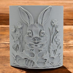 以下が含まれることがあります： 蝶ネクタイをした灰色の3Dプリントされたイースターバニー。花と卵に囲まれています。