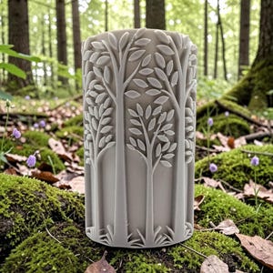 Può includere: Un portacandele cilindrico grigio chiaro con un rilievo dettagliato di alberi e foglie stilizzate. Il portacandele è ambientato in un ambiente forestale con muschio verde, foglie cadute e fiori selvatici viola.