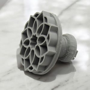 May include: A gray plastic cookie cutter shaped like a flower with a detailed design. The cutter has a handle on the back.