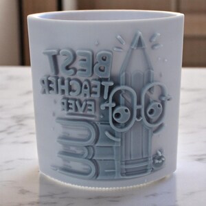 May include: A light blue ceramic mug with the words "BEST TEACHER EVER" embossed on the front. The design includes a pencil, books, and glasses. The mug is cylindrical and sits on a light-colored surface.
