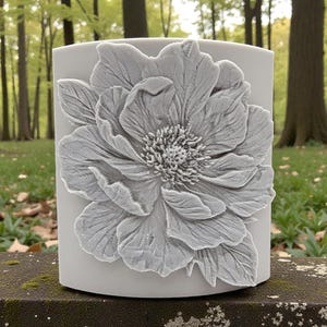 May include: A cylindrical light gray vessel featuring a prominent, three-dimensional relief of a large, multi-petaled flower on its front surface. The flower design displays intricate textures and layered petals, creating a tactile appearance.