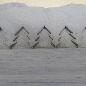 May include: A close-up of a gray clay surface with five embossed Christmas tree designs. The trees are stylized and have a simple, geometric design.