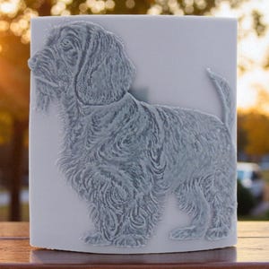 May include: A light gray, rectangular decorative tile featuring a detailed relief of a dog, likely a Wirehaired Pointing Griffon. The dog is depicted in profile, with textured fur and a bushy tail. The background is blurred with warm sunlight.