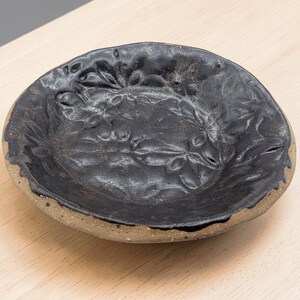 Op de afbeelding: Een handgemaakte, ovale keramische kom met een donker, getextureerd interieur en een lichtere, gespikkelde buitenkant. De kom heeft een reliëf bloemenontwerp aan de binnenkant. De rand is zwart geschilderd en de kom heeft een diameter van ongeveer 15 cm.