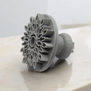 May include: A gray, 3D-printed cookie cutter in the shape of a flower. The cutter has a detailed floral design on the cutting edge and a handle for easy use. The cutter is approximately 3 inches in diameter.